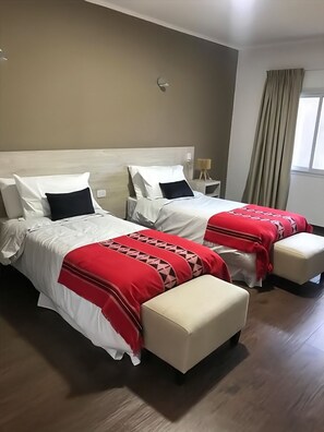 A hotel room with two beds, each with white bedding and red accents, a beige wall, a window with curtains, and a wooden floor.
