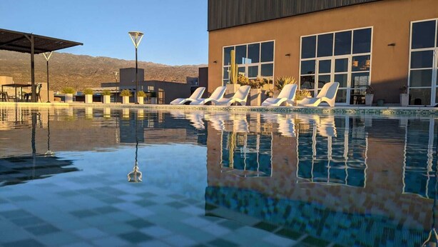 A swimming pool with lounge chairs, a lamp post, and a building with large windows and plants. - ENTRE VIÑEDOS RESORT HOTEL  (Schaqui)