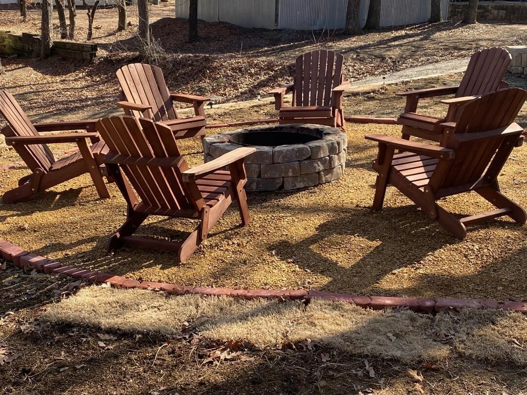 A fire pit surrounded by six Adirondack chairs in a backyard setting.