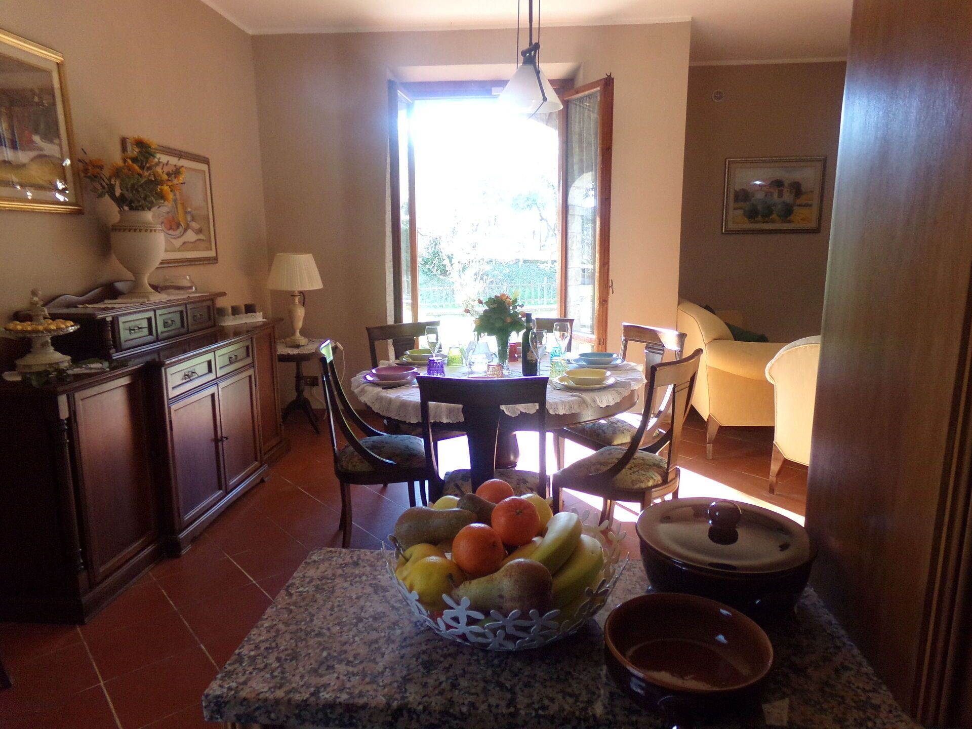 Un coin repas avec une table dressée pour deux personnes, un panier de fruits sur la table et une vue sur l’extérieur par une fenêtre.