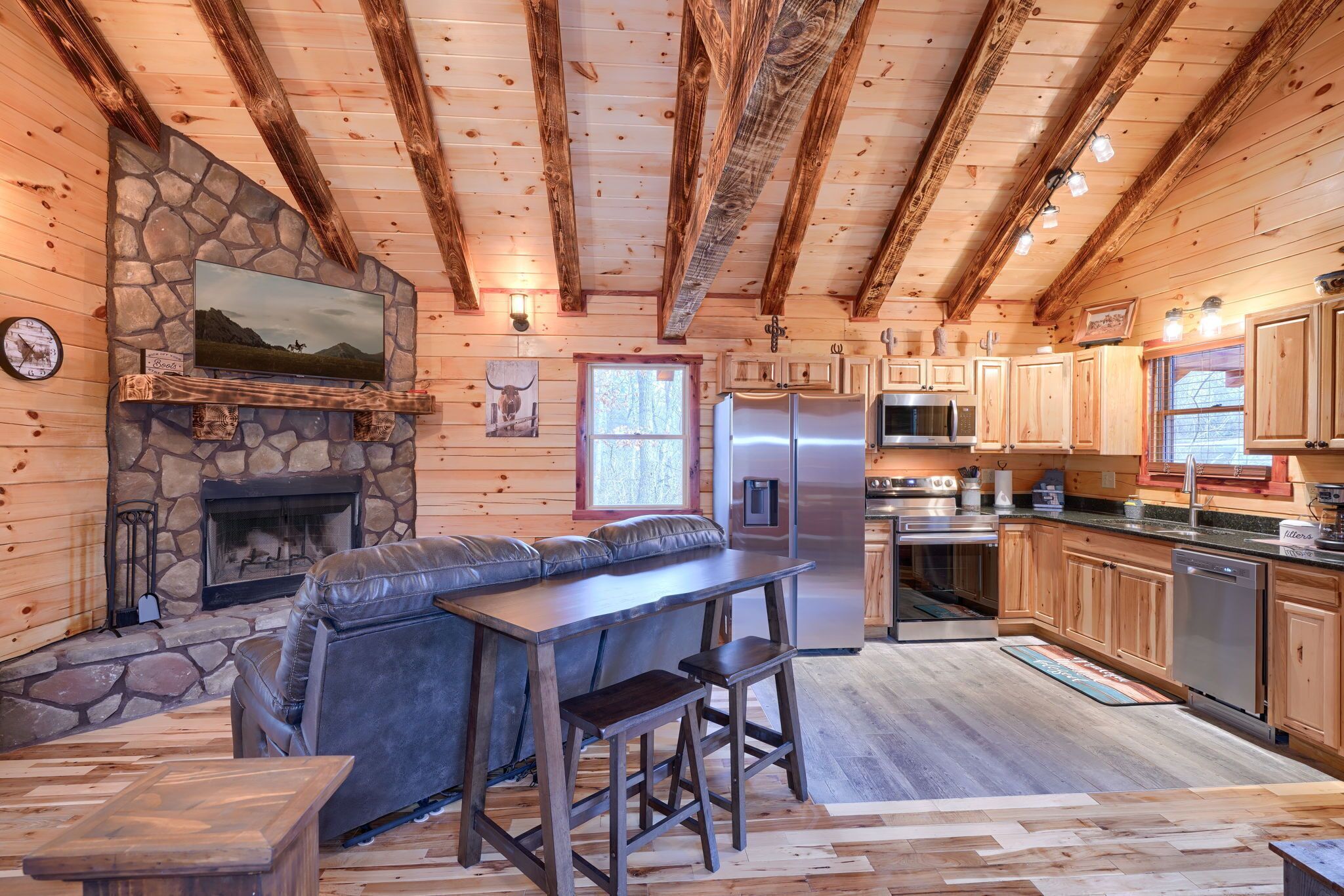 A cozy cabin interior with a stone fireplace, a stone wall, a television mounted above it, a wooden ceiling with exposed beams, a kitchen area with wooden cabinets, a refrigerator, a microwave, and a dining area with a bench and stools.