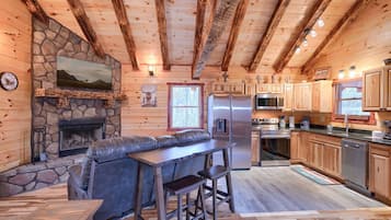 A cozy cabin interior with a stone fireplace, a stone wall, a television mounted above it, a wooden ceiling with exposed beams, a kitchen area with wooden cabinets, a refrigerator, a microwave, and a dining area with a bench and stools.