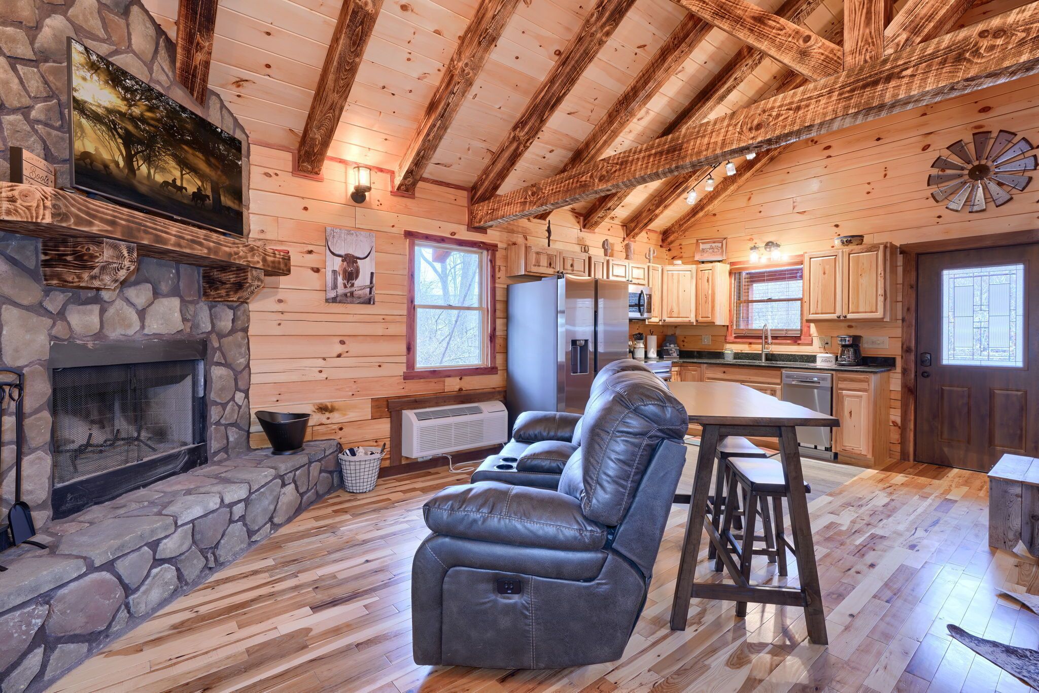 A cozy cabin interior with a stone fireplace, leather recliner, wooden ceiling beams, and a kitchen area.