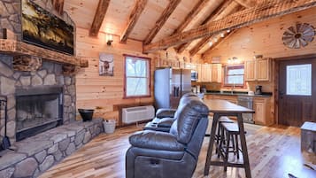 A cozy cabin interior with a stone fireplace, leather recliner, wooden ceiling beams, and a kitchen area.