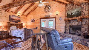 Interior de uma cabana de toras com uma cama, lareira de pedra, área de bar e uma pintura pendurada na parede.