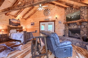 A log cabin interior with a stone fireplace, a bar area, a bed with a cowhide rug, and a welcome sign.