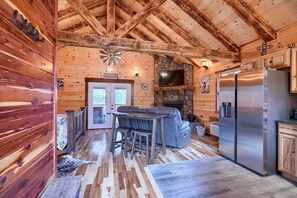 A log cabin interior with a stone fireplace, wooden ceiling beams, a bar area, and a modern kitchen with stainless steel appliances. - Cowboy Trail - Lovely Cabin for 2 in Hocking Hills (Logan)