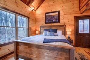 Un dormitorio de cabaña de madera con una cama grande, un cuadro enmarcado sobre la cama y una ventana con vistas a árboles.