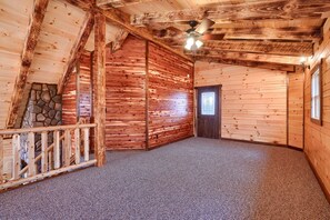 Interior de una cabaña de troncos con chimenea de piedra, paredes de madera, ventilador de techo y piso alfombrado.