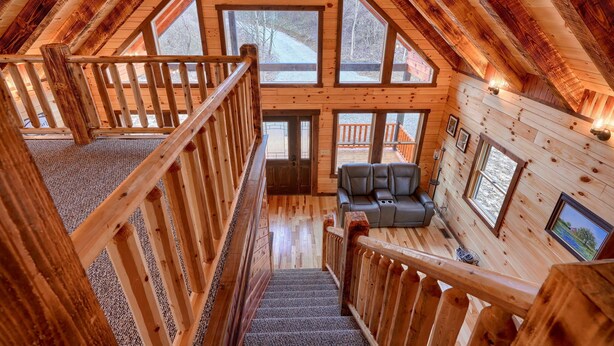 A wooden interior with a staircase leading to a loft area, a seating area with a sofa, and large windows offering views of the outdoors. - Bluebonnet Luxury Cabin - in the Hocking Hills (Logan)