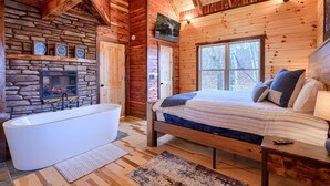A log cabin interior with a stone fireplace, a large bathtub, and a bed with a view of the outdoors. - Bluebonnet Luxury Cabin - in the Hocking Hills (Logan)
