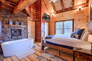 Un dormitorio de cabaña de madera con chimenea de piedra, un ventanal grande, una cama con almohadas y una bañera.