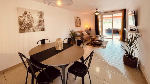 Un coin repas avec une table ronde, des chaises et un luminaire suspendu. On aperçoit un canapé et un téléviseur à l’arrière-plan.
