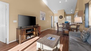 A spacious living room with a flat-screen TV, wooden entertainment unit, and a dining area with a table and chairs.