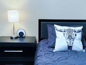 A neatly made bed with a patterned pillow, a bedside lamp, and a small table with a lamp and a clock.
