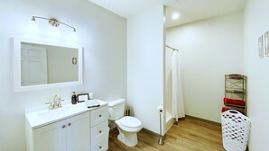 Ein Badezimmer mit weißem Waschbecken, Spiegel und Toilette. Handtuchhalter und Wäschekorb sind ebenfalls vorhanden.