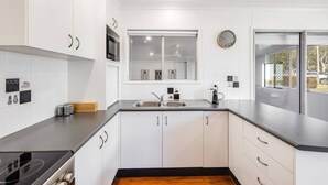 A modern kitchen with white cabinetry, a black countertop, a built-in stove, a double sink, and a window with a clock. - "Eastside Haven" - Central & Convenient! (East Toowoomba)