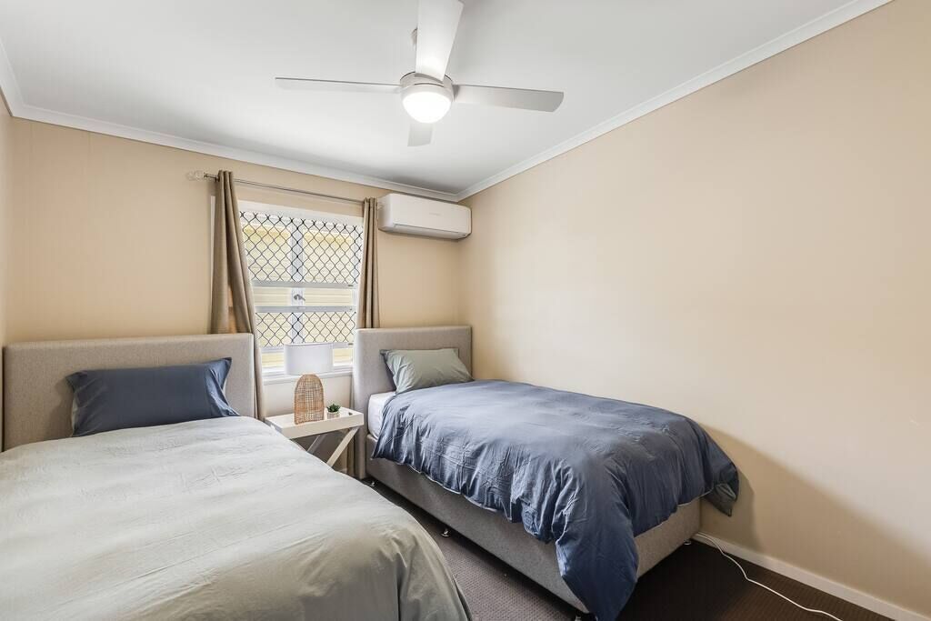 A room with two beds, a ceiling fan, an air conditioning unit, and a window with a metal grille.