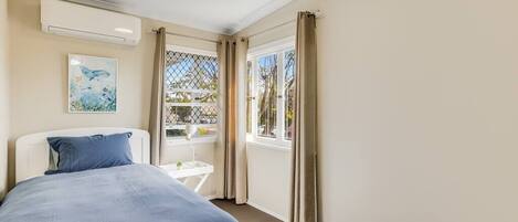 A bedroom with a bed, a window with curtains, a small table, and a wall-mounted air conditioner.