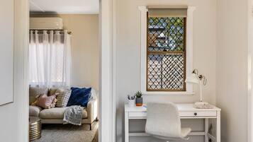 A room with a white desk, a chair, a window with a decorative screen, a sofa with cushions, and a radiator.