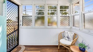 A sunroom with a wicker chair, a small table with a plant, and a view of the outside through frosted windows. - Cheerful, Cosy Cottage - Near Rose Garden! (Newtown)