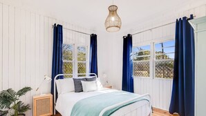 A bedroom with a white metal bed, a bedside table with a lamp, a potted plant, and a window with blue curtains. - Cheerful, Cosy Cottage - Near Rose Garden! (Newtown)