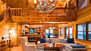 A log cabin interior with a cozy living area, wooden walls, a chandelier, and a view through the windows. - Summit-Level Luxury: Views, Hot Tub, Games and 15 minutes to town! (Blue Ridge)