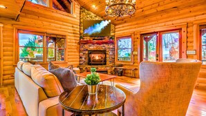 A log cabin interior with a stone fireplace, wooden walls, a chandelier, and a large window overlooking a forest. - Summit-Level Luxury: Views, Hot Tub, Games and 15 minutes to town! (Blue Ridge)