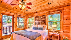 A log cabin bedroom with a large bed, wooden walls, and multiple windows offering views of the surrounding forest. - Summit-Level Luxury: Views, Hot Tub, Games and 15 minutes to town! (Blue Ridge)