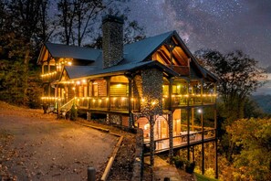 A log cabin with a stone chimney, multiple decks, and string lights.