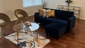 Een woonkamer met een donkerblauwe bank, twee gouden fauteuils, een glazen salontafel en een vloerkleed met dessin.