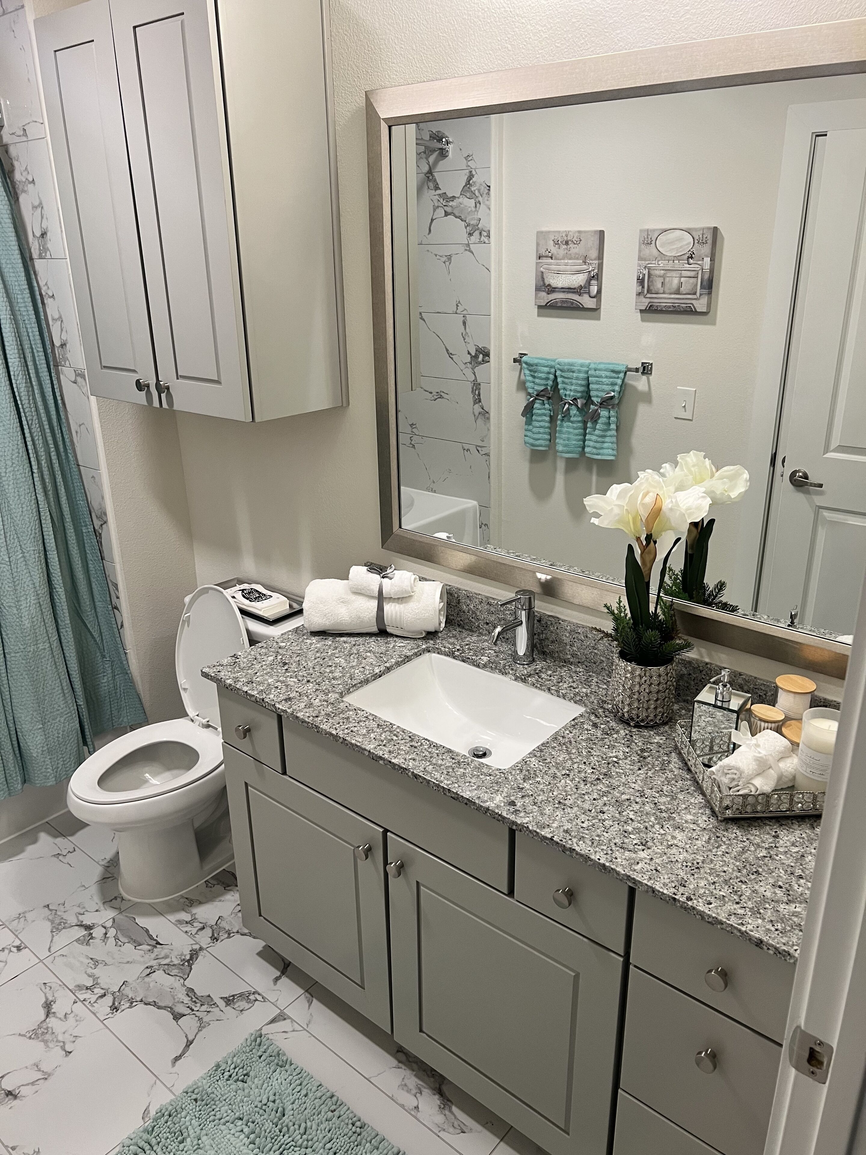 A bathroom with a white sink, granite countertop, and a mirror above it. There is a toilet to the left and a towel rack with towels above it.
