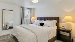 A neatly made bed with white linens and a dark headboard, two bedside lamps, a large mirror, and a window with curtains. - Sunset Paradise Retreat | 2 Hot Tubs | 2 Houses (Orem)