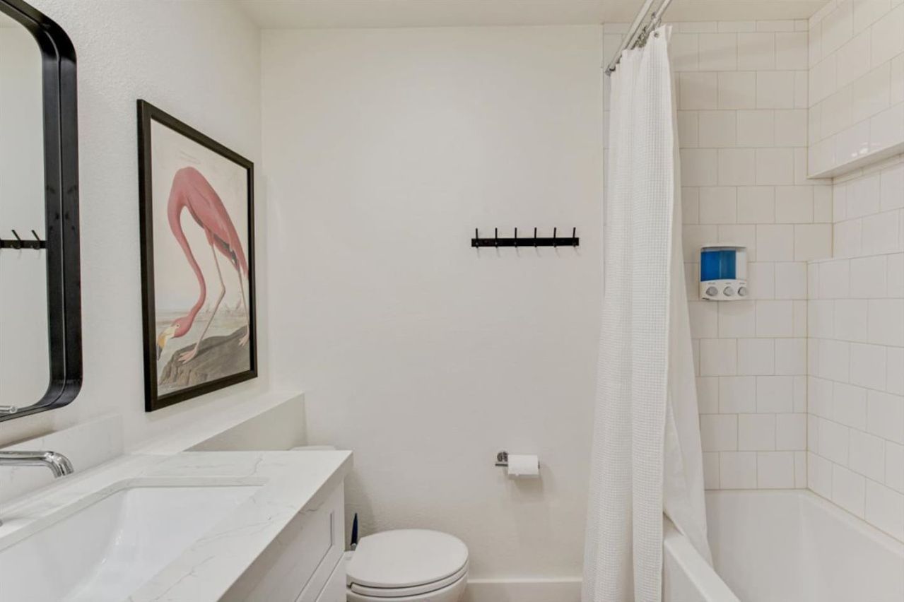 Un baño con inodoro blanco, lavamanos, espejo, un cuadro enmarcado, cortina de ducha y dispensador de jabón.