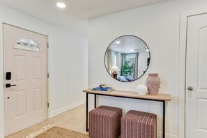 A hallway with a white door, a round mirror, a small table with a lamp, and two striped ottomans.