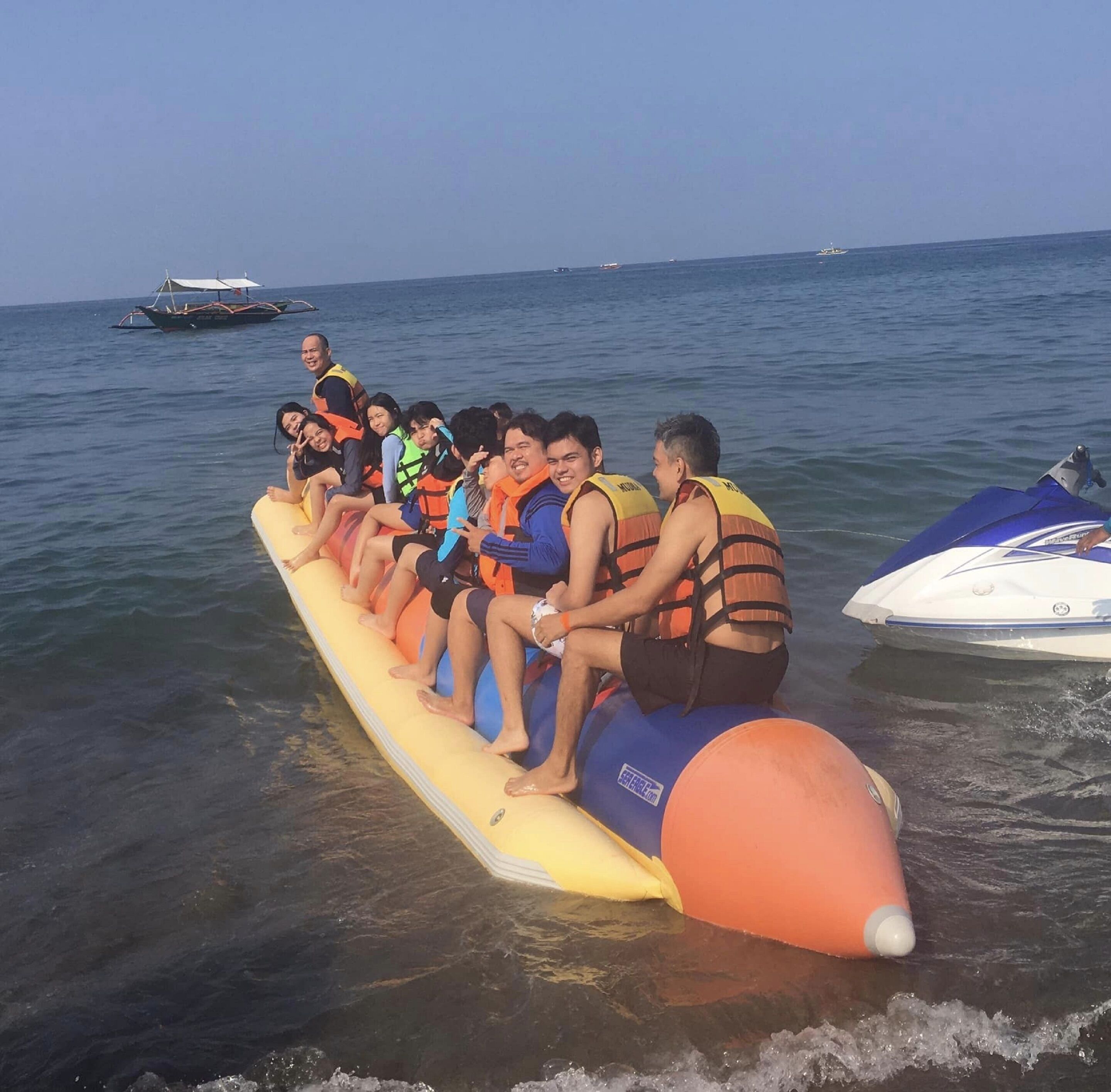A group of people on a watercraft with life jackets, a boat in the background, and a jet ski to the right.