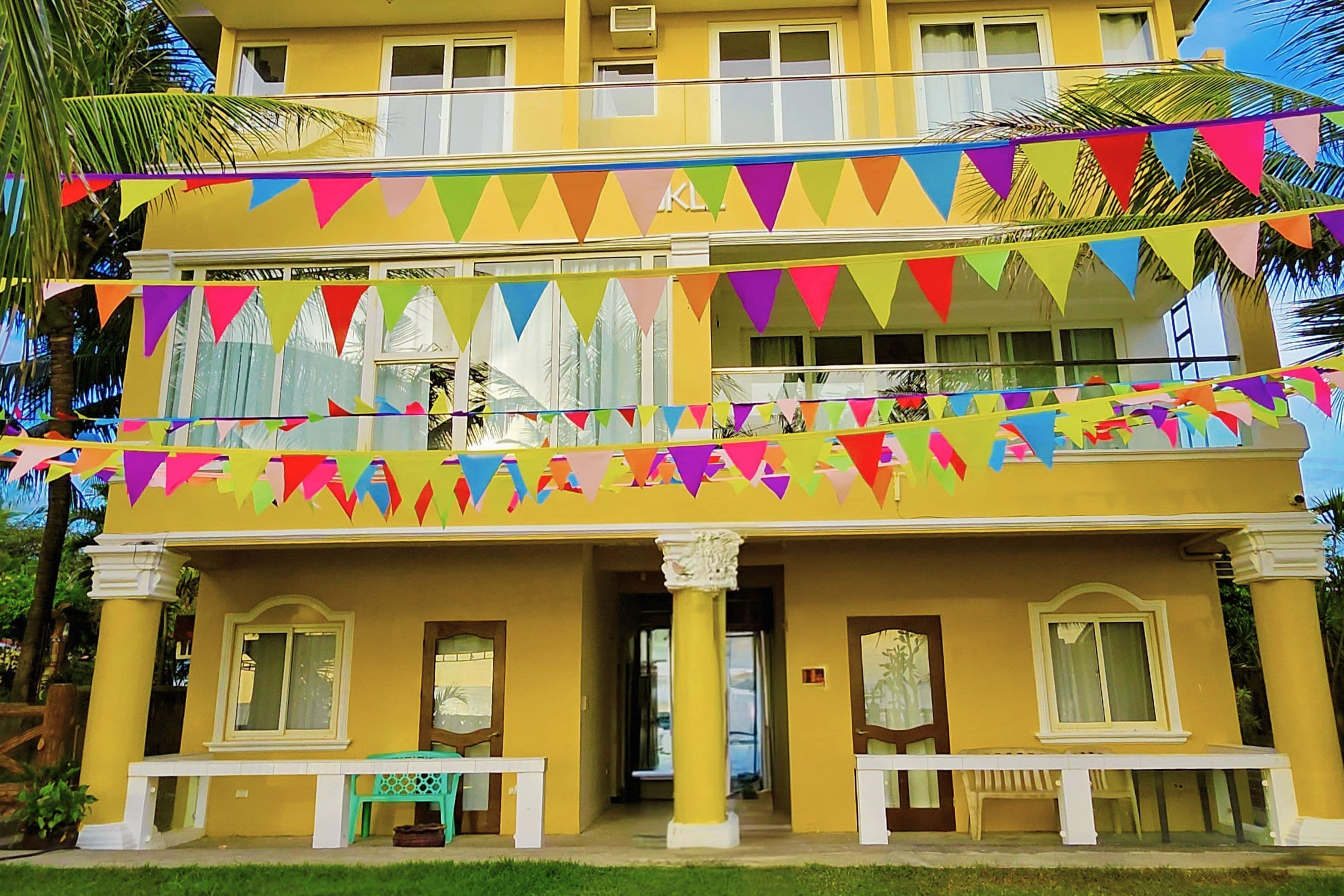 A two-story building with a yellow facade, balconies, and colorful triangular flags.
