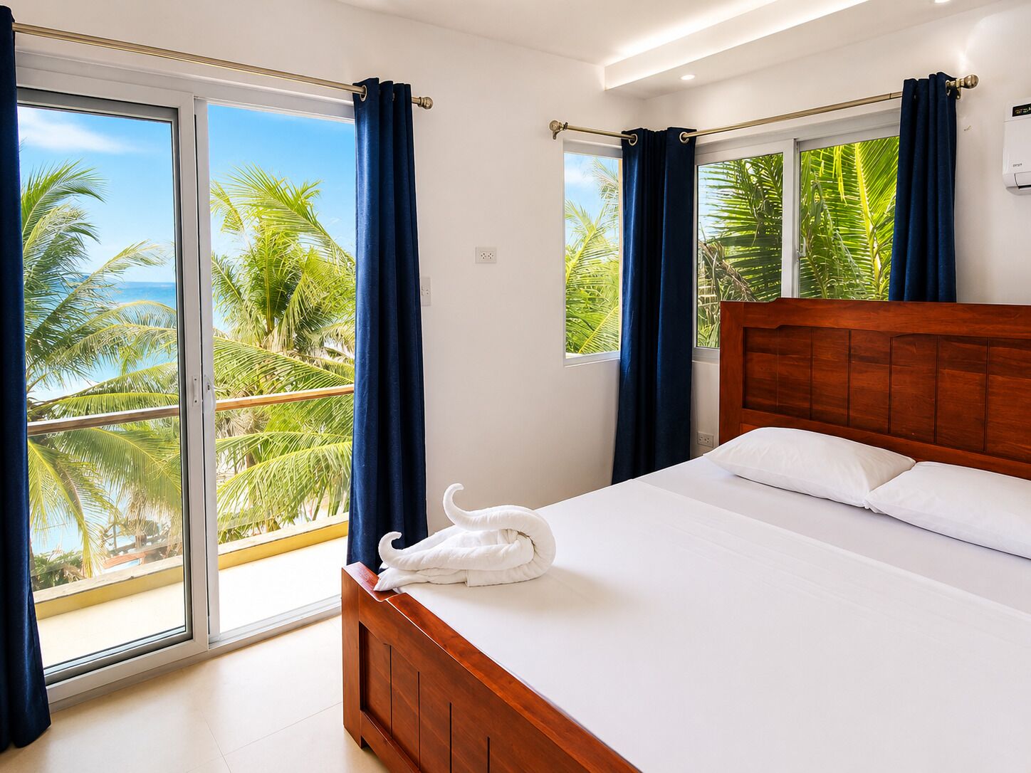 Quarto com cama de madeira, vista para palmeiras e oceano através de grandes janelas, e uma toalha sobre a cama.