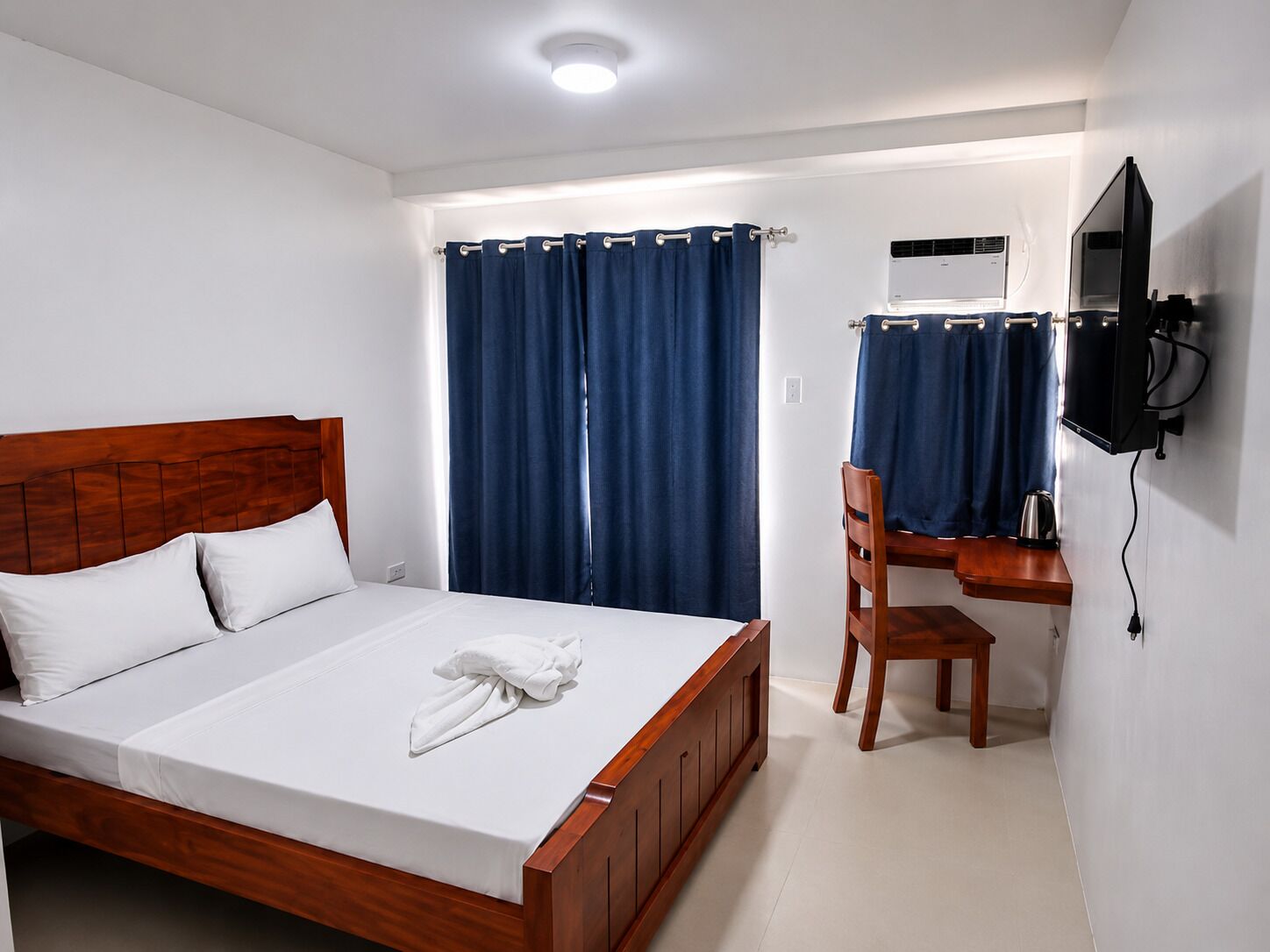 Habitación de hotel con cama de madera, un escritorio con silla, televisión y ventana con cortinas.