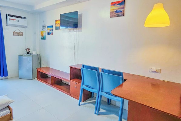 A room with a wooden dining table, blue chairs, a red bench, a small refrigerator, and a TV mounted on the wall. - Crystal Shore Beach Resort powered by Cocotel (Morong)