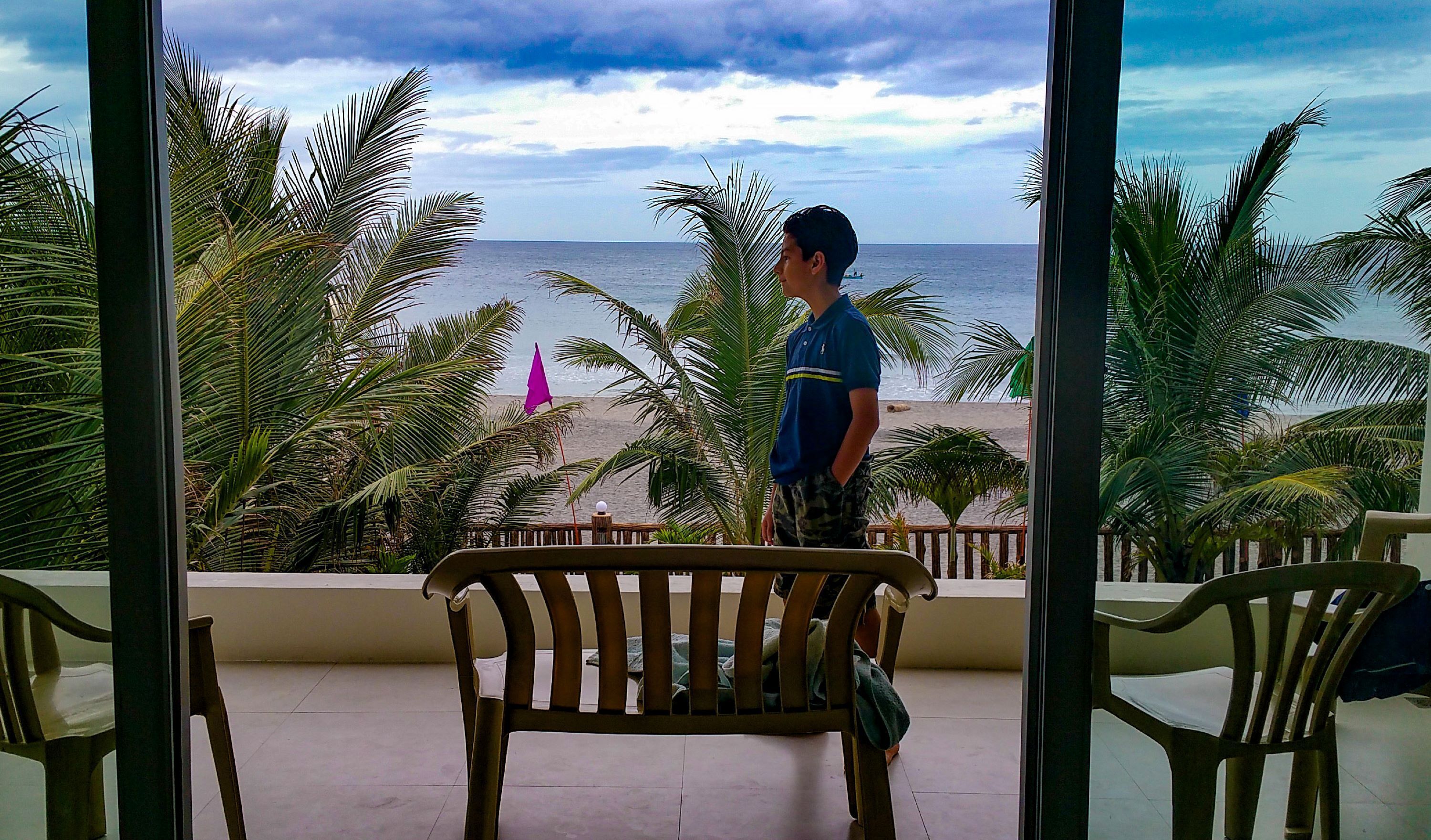 Uma varanda com vista para a praia, palmeiras e uma pessoa em pé.