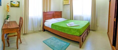 A neatly arranged bedroom with a wooden bed, a green bedspread, a wooden nightstand, a chair, and a small table with a vase of flowers.