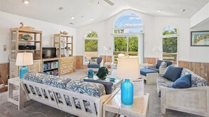 A spacious living room with a wooden sofa, a coffee table, and a TV. There are bookshelves and a ceiling fan. - Casa Sonada II - 4 BR Beach Home in Quiet, Low Traffic Neighborhood, Dogs Allowed (Southern Shores)