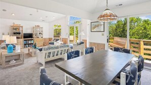 A spacious living room with a large table and chairs, a sofa, and a TV. There are wooden shelves with books and decorative items, and a large window offering a view of greenery outside. - Casa Sonada II - 4 BR Beach Home in Quiet, Low Traffic Neighborhood, Dogs Allowed (Southern Shores)