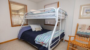Habitación con literas, una mecedora de madera, estructura metálica blanca y ventana con persianas.