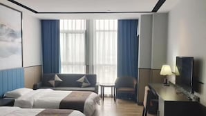 A hotel room with two beds, a sofa, a desk with a lamp, a TV, and large windows with curtains.