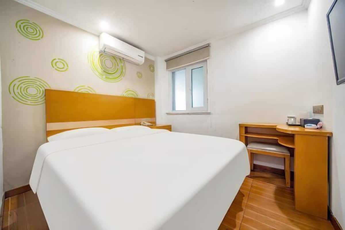 A hotel room with a bed, a desk, a chair, a window, and a wall-mounted air conditioner.