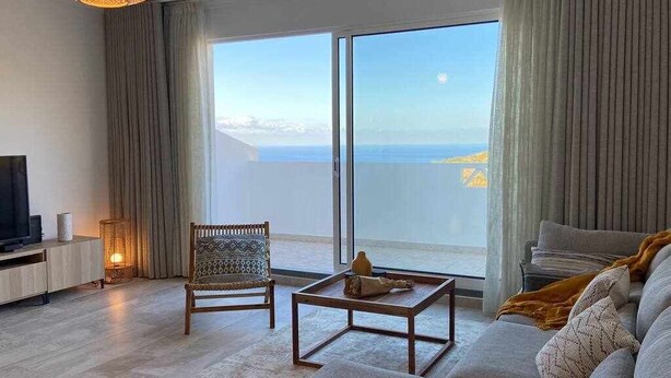 A modern living room with a large sliding glass door leading to a balcony with a sea view. - Garajau Apart with Amazing Views, Parking & Wifi. (Caniço)