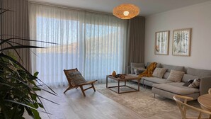 A modern living room with a sofa, a wooden chair, a coffee table, and a large window with sheer curtains. - Garajau Apart with Amazing Views, Parking & Wifi. (Caniço)