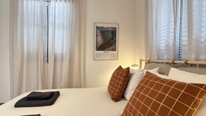 Habitación de hotel con cama, cortinas blancas, un cuadro en la pared y una mesita de noche con lámpara.